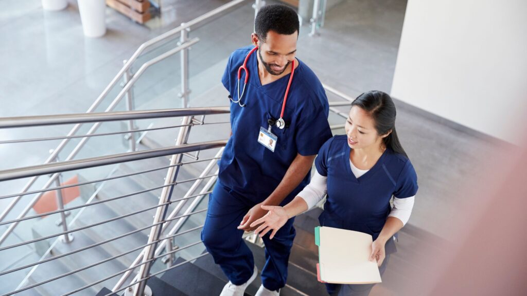 Two nurses in scrubs walking up a staircase. One holding a clipboard with paperwork.
